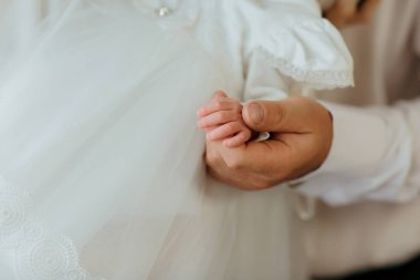 Beyaz elbiseli biri tarafından bir bebek tutuluyor. Bebek beyaz bir elbise giyiyor ve insan bebeğin elini tutuyor.