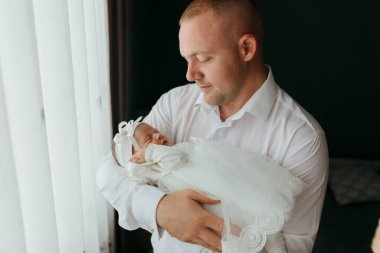 Bir adam kucağında bir bebek tutuyor. Bebek beyaz bir elbise ve beyaz bir saç bandı takıyor. Adam gülümsüyor ve bebek uyuyor. Erkek ve bebek arasındaki sıcaklık ve sevgi kavramı.