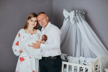 Genç bir çift beşikte bir bebek tutuyor. Bebek beyaz bir elbise giyiyor. Oda beyaz bir gölgelikle süslenmiş.