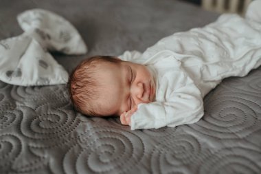 Bir bebek battaniyeli ve şapkalı bir yatakta uyuyor. Bebek beyaz bir tulum giyiyor.