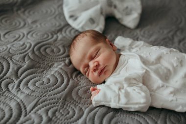 Bir bebek üzerinde beyaz bir fiyonk olan bir yatakta uyuyor. Bebek beyaz bir tulum giyiyor ve yüzünde bir el var. Sahne huzurlu ve rahatlatıcı, bebeğin yumuşak nefesiyle.