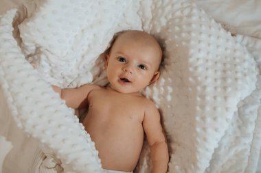 Bir bebek beyaz bir battaniyenin üzerine uzanmış başını yana çevirmiş yatıyor. Bebek gülümsüyor ve mutlu.