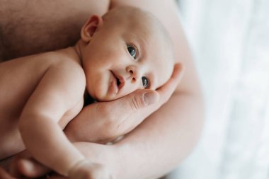 Bir bebek, gözleri kapalı ve yüzünde bir gülümsemeyle bir ebeveyn tarafından tutuluyor. Bebek bir battaniyeye sarılmış ve küçük parmakları ebeveynin eline dolanmış.