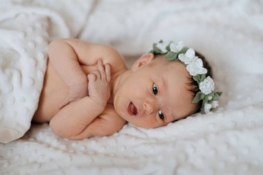 Çiçekli saç bandı takan bir bebek beyaz bir battaniyeye uzanır. Bebek gülümsüyor ve kameraya bakıyor.