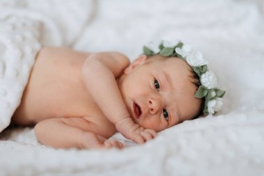 Çiçekli saç bandı takan bir bebek beyaz bir battaniyeye uzanır. Bebek kameraya bakıyor.