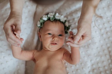 Çiçekli saç bandı takan bir bebek iki kişi tarafından tutuluyor. Bebek gülümsüyor ve kameraya bakıyor.