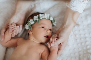 Çiçekli saç bandı takan bir bebek iki kişi tarafından tutuluyor. Bebek beyaz bir battaniyenin üzerinde yatıyor.