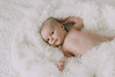 Beyaz bir battaniyenin üzerinde bir bebek yatıyor. Bebek gülümsüyor ve mavi gözlü.