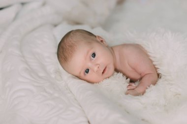 Bir bebek gözleri açık beyaz bir battaniyenin üzerinde yatıyor. Bebek kameraya bakıyor, sahne huzurlu ve huzurlu.