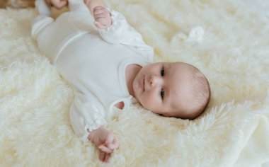 Beyaz bir battaniyenin üzerinde bir bebek yatıyor. Bebek beyaz bir elbise giyiyor ve üzerinde beyaz bir battaniye var.