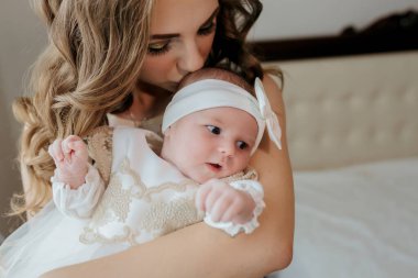Bir kadın kucağında bir bebek tutuyor. Bebek beyaz bir saç bandı ve beyaz bir elbise giyiyor. Kadın bebeğin alnını öpüyor. Anne ve çocuğu arasındaki sıcaklık ve sevgi kavramı.