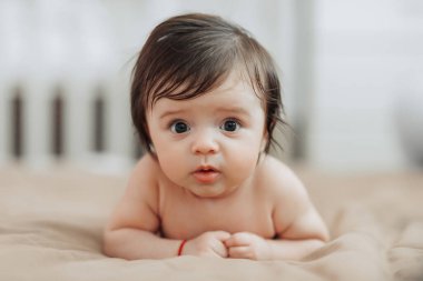 Bir bebek gözleri açık bir şekilde bir yatağın üzerinde yatıyor. Bebeğin yüzü görüntünün ana odak noktasıdır ve merak uyandırır ya da şaşırtır. Yatak kahverengi bir battaniyeyle kaplı.