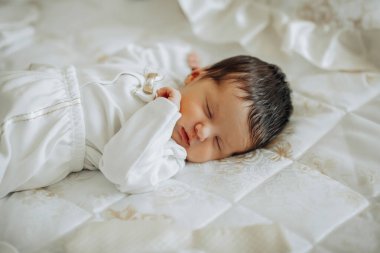 Bir bebek bir yatakta uyuyor. Bebek beyaz bir kıyafet giyiyor. Yatak beyaz bir battaniyeyle kaplı.