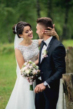 Bir gelin ve damat tahta bir çitin yanında kameraya gülümsüyor. Gelin bir buket çiçek tutuyor ve damat bir takım elbise giyiyor. Sahne mutlu ve kutlama dolu.