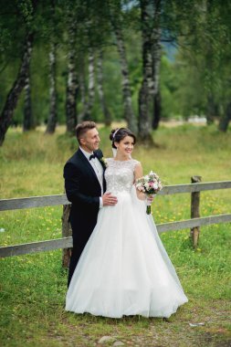 Bir gelin ve damat, beyaz bir elbise ve siyah bir takım elbiseyle bir tarlada duruyor. Gelin elinde bir buket çiçek tutuyor.