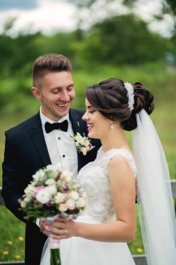 Bir gelin ve damat kameraya gülümsüyor. Gelin beyaz bir elbise ve duvak takıyor. Damat siyah bir takım elbise ve papyon takıyor. Bir buket ve yaka çiçeği tutuyorlar.