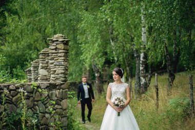 Gelin ve damat bir ormanda, gelin bir buket tutuyor. Sahne romantik ve huzurlu, çünkü çift doğa ile çevrili.