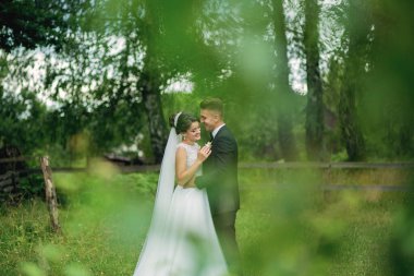 Bir gelin ve damat arka planda ağaçlarla bir tarlada duruyorlar. Gülümsüyorlar ve el ele tutuşuyorlar.