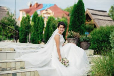 Beyaz elbiseli bir kadın, kucağında bir buket çiçekle merdivenlerde oturuyor.