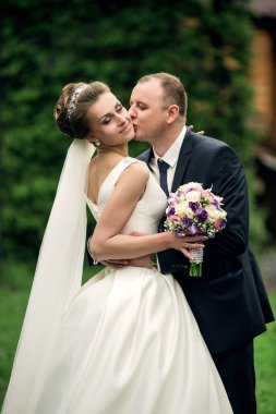 Bir gelin ve damat birbirlerine sarılıp öpüşüyorlar. Gelin beyaz bir elbise giyiyor ve damat bir takım elbise giyiyor. Bir buket ve vazo tutuyorlar.