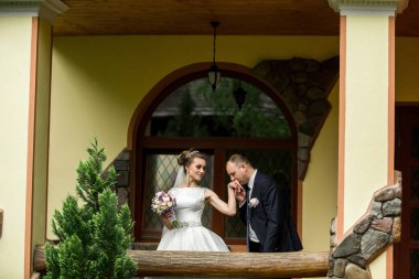 Gelin ve damat balkonda duruyorlar, gelin buketi tutuyor ve damat elini tutuyor.