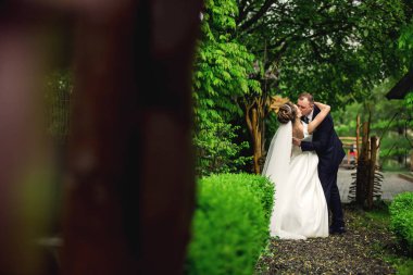 Bir gelin ve damat yemyeşil bir ormanda öpüşüyor. Gelin beyaz bir elbise giyiyor ve damat bir takım elbise giyiyor. Sahne romantik ve mutlu.