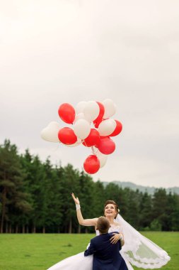 Bir gelin ve damat bir demet kırmızı ve beyaz balon tutarken, gelin adam tarafından havaya kaldırılıyor. Çiftin etrafı renkli balonlarla çevriliyken, neşe ve kutlama kavramı