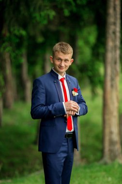 Mavi takım elbiseli ve kırmızı kravatlı genç bir adam yeşil bir ormanda duruyor. Kırmızı bir kravat takıyor ve cebinde kırmızı bir gül var.