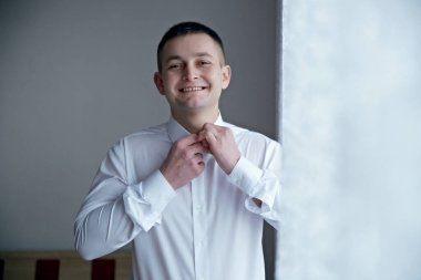 Beyaz gömlekli bir adam giyiniyor ve gülümsüyor. Beyaz bir gömlek ve kravat takıyor.