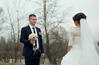 Bir gelin ve damat bir buket ve kravatla bir tarlada duruyor. Gelin beyaz bir elbise giyiyor ve damat bir takım elbise giyiyor. Sahne romantik ve mutlu.