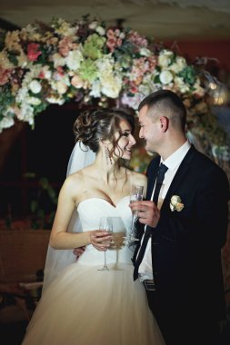 Bir gelin ve damat elinde şampanya kadehleriyle çiçek kemerinin önünde duruyorlar. Gülümsüyorlar ve birbirlerine bakıyorlar, anın tadını çıkarıyorlar.