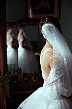 Gelinlikli bir kadın aynanın önünde duruyor. Duvak ve beyaz bir elbise giyiyor.