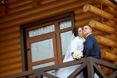 Gelin ve damat ahşap bir balkonda duruyorlar, gelin de elinde buketle. Sahne romantik ve samimi, çünkü çift birlikte poz veriyor.