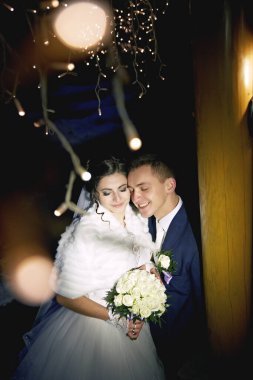 Bir gelin ve damat bir buket çiçekli fotoğraf için poz veriyorlar. Gelin beyaz kürk şort giyiyor ve damat mavi bir takım giyiyor. Sahne mutlu ve romantik.