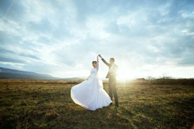 Bir gelin ve damat, üzerinde güneş olan bir tarlada dans ediyorlar. Sahne neşeli ve romantik.