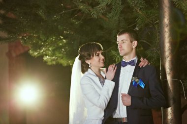 Gelin ve damat bir ağacın altında, gelin beyaz bir elbise ve damat mavi bir takım elbise giyiyor.