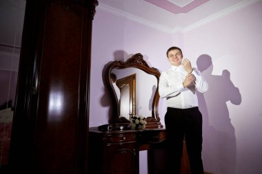 Bir adam aynanın önünde duruyor, giyiniyor. Oda loş bir şekilde aydınlatılmış ve adamın yansıması aynada görülüyor.