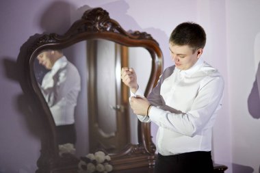 Bir adam aynanın önünde giyiniyor. Beyaz gömlek ve siyah pantolon giyiyor.