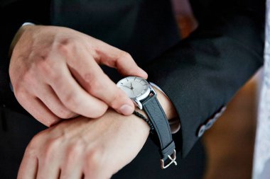 Bir adam saat takıyor ve ona bakıyor. Saat siyah ve gümüş. Adam siyah takım elbise ve beyaz gömlek giyiyor.