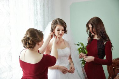 Üç kadın düğün için hazırlanıyor. Biri gelinlik giyiyor ve diğer ikisi de ona yardım ediyor.