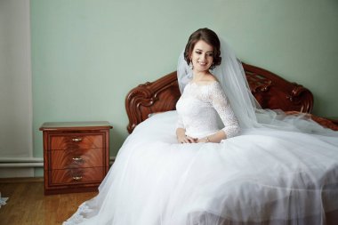 Bir gelin, odasındaki bir yatakta ahşap bir şifoniyerle oturuyor. Beyaz bir gelinlik ve duvak giyiyor.