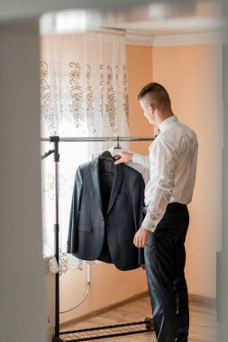 Bir adam asılmış bir ceketin önünde kravatını düzeltiyor. Formalite ve detaylara dikkat etme kavramı. Adam özel bir olaya ya da önemli bir olaya hazırlanıyor.