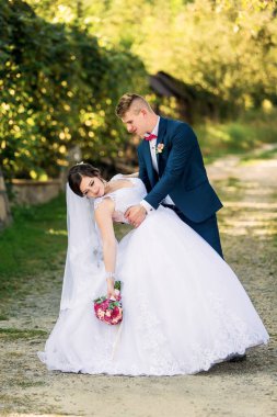 Bir gelin ve damat toprak yolda fotoğraf çektirmek için poz veriyor. Gelin beyaz bir elbise giyiyor ve elinde bir buket çiçek tutuyor. Damat takım elbise ve kırmızı kravat takıyor. Sahne romantik ve mutlu.