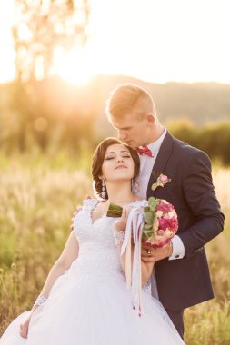 Bir gelin ve damat güneş üzerlerine doğarken bir tarlada dikiliyorlar. Gelin bir buket tutuyor ve damat papyon takıyor. Sahne mutlu ve romantik.