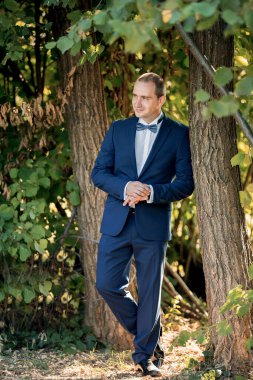 Takım elbiseli ve papyonlu bir adam bir ağacın önünde duruyor. Mavi bir takım ve beyaz bir gömlek giyiyor.