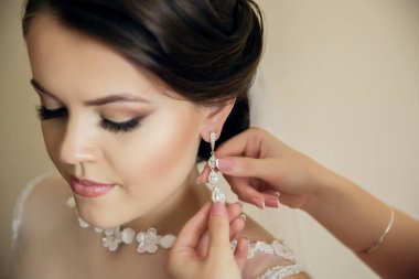 Bir kadın küpelerini bir kadına taktırıyor. Kadın beyaz bir elbise giyiyor ve saçları topuz şeklinde.