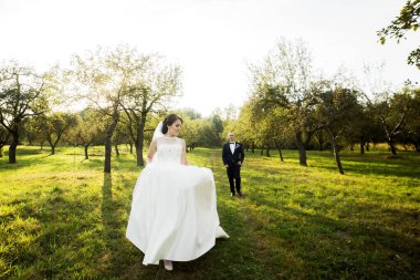 Bir gelin ve damat ağaçların arasında yürüyor. Gelin beyaz bir elbise giyiyor ve damat bir takım elbise giyiyor. Sahne huzurlu ve romantik.