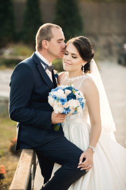 Bir gelin ve damat bir buket çiçek tutarken birbirlerini öpüyorlar. Gelin beyaz bir elbise giyiyor ve damat mavi bir takım elbise giyiyor. Sahne romantik ve mutlu.