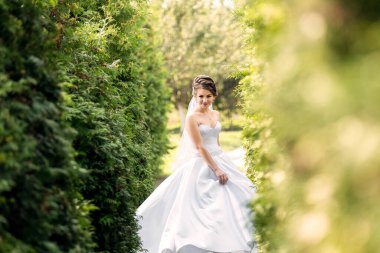 Beyaz bir elbise giymiş bir gelin yeşil bir çitin içinden geçiyor. Gülümsüyor ve kameraya bakıyor.