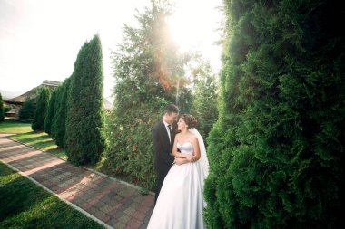 Gelin ve damat bir çitin önünde duruyorlar, gelin beyaz bir elbise giyiyor ve damat takım elbise giyiyor.
